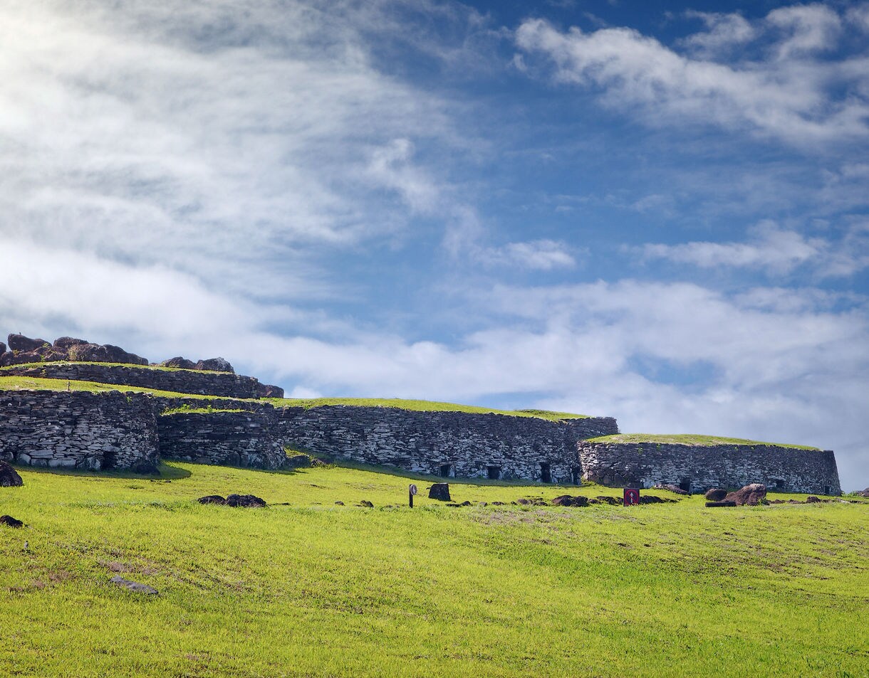 Low stone structures of Orongo Village on a grassy hillside under a wide blue sky with scattered clouds on Easter Island.