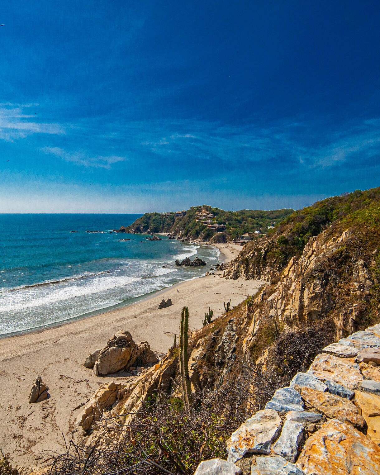 Rugged coastline at Copalita eco-archaeological park with golden sandy beach, rocky cliffs and waves rolling in under a deep blue sky.