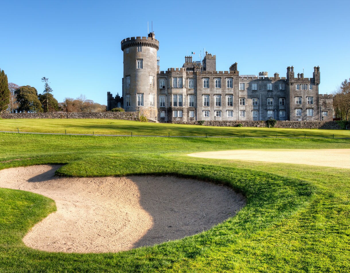 Stone castle with a round tower rising behind a golf course bunker and manicured fairway.