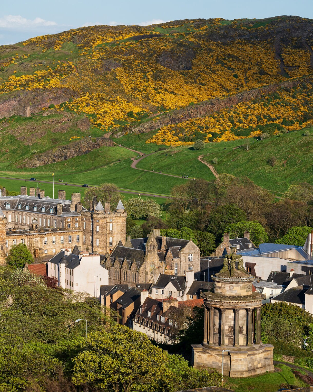 Holyrood Palace in Edinburgh surrounded by lush trees and gardens, with Arthur’s Seat rising behind in green slopes dotted with yellow gorse.