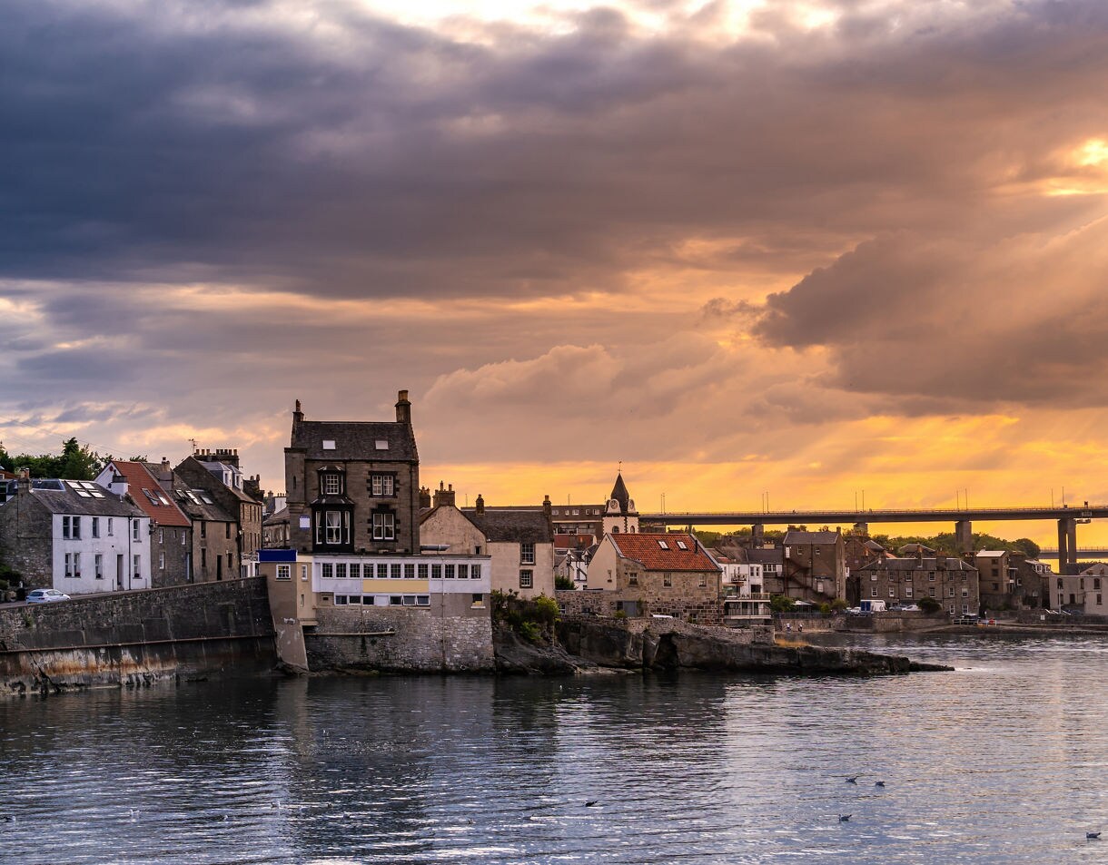 Coastal village of Queensferry with stone houses and a bridge in the background at sunset.
