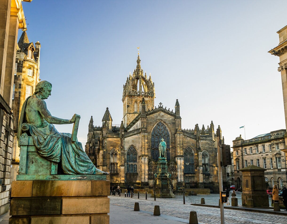 View of St. Giles’ Cathedral in Edinburgh with a bronze statue of David Hume in the foreground and cobblestone streets surrounding the Gothic church.
