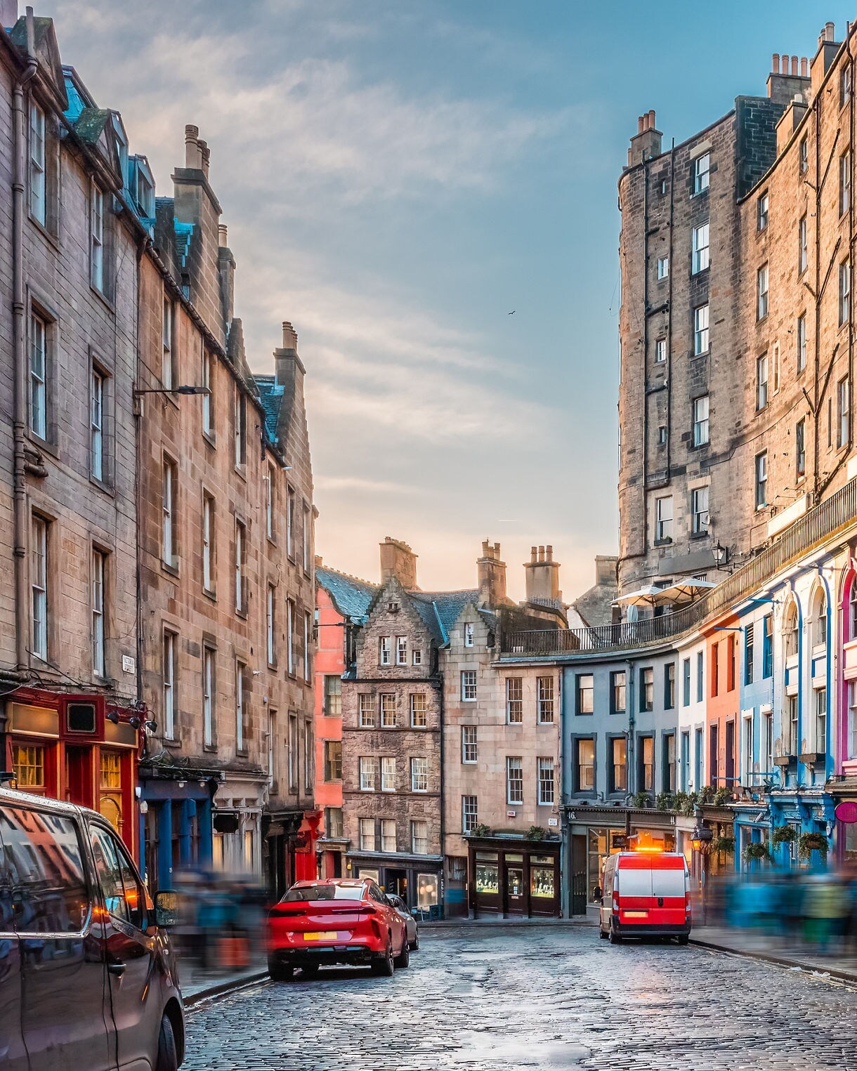 Curved cobblestone Victoria Street in Edinburgh lined with tall stone buildings and brightly painted storefronts in red, blue and purple.