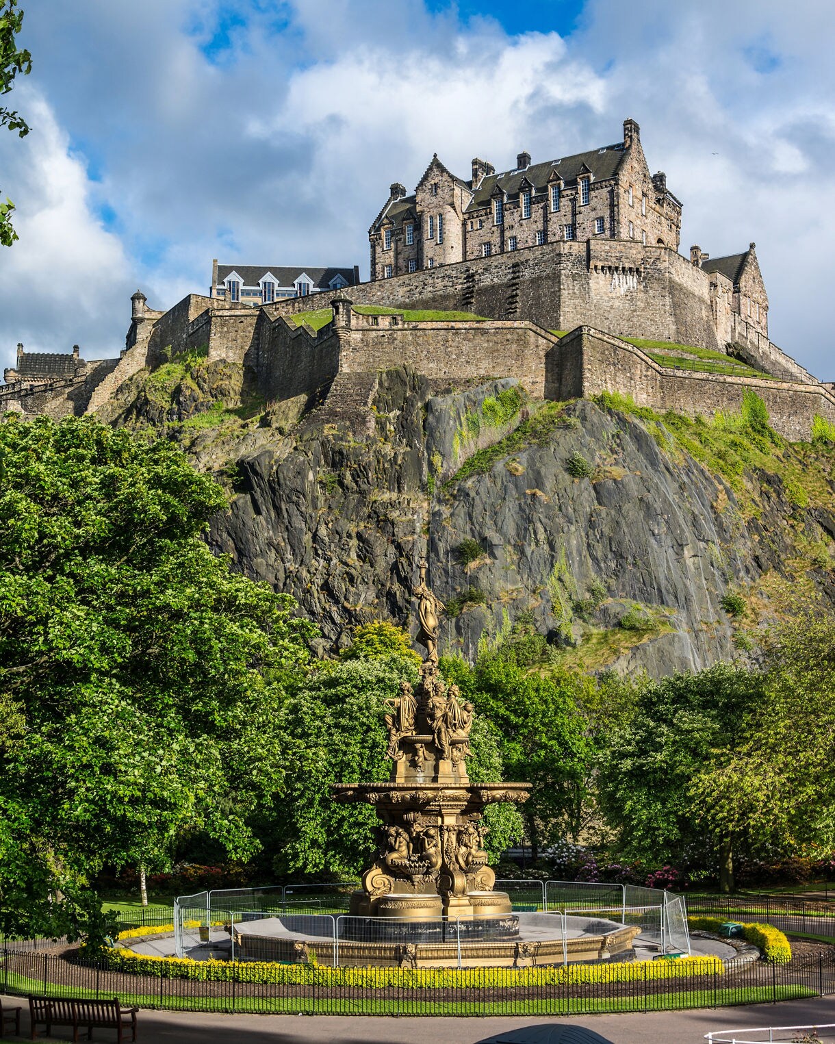 Edinburgh Castle on a rocky cliff with a decorative fountain and trees in the foreground.