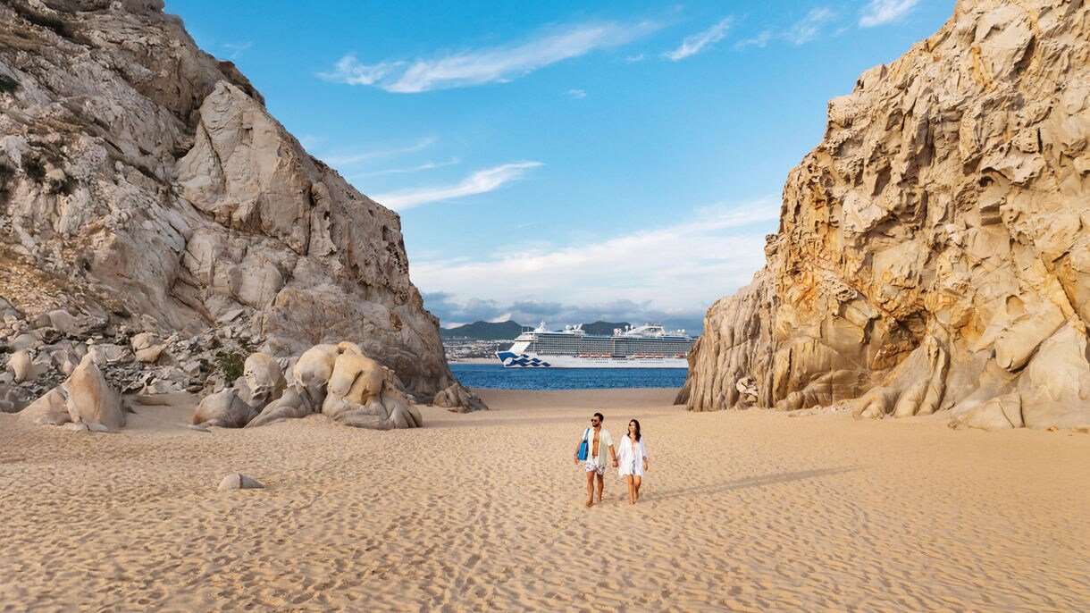 Two people walk on a sandy beach between dramatic golden rock formations with a cruise ship visible in the distance.