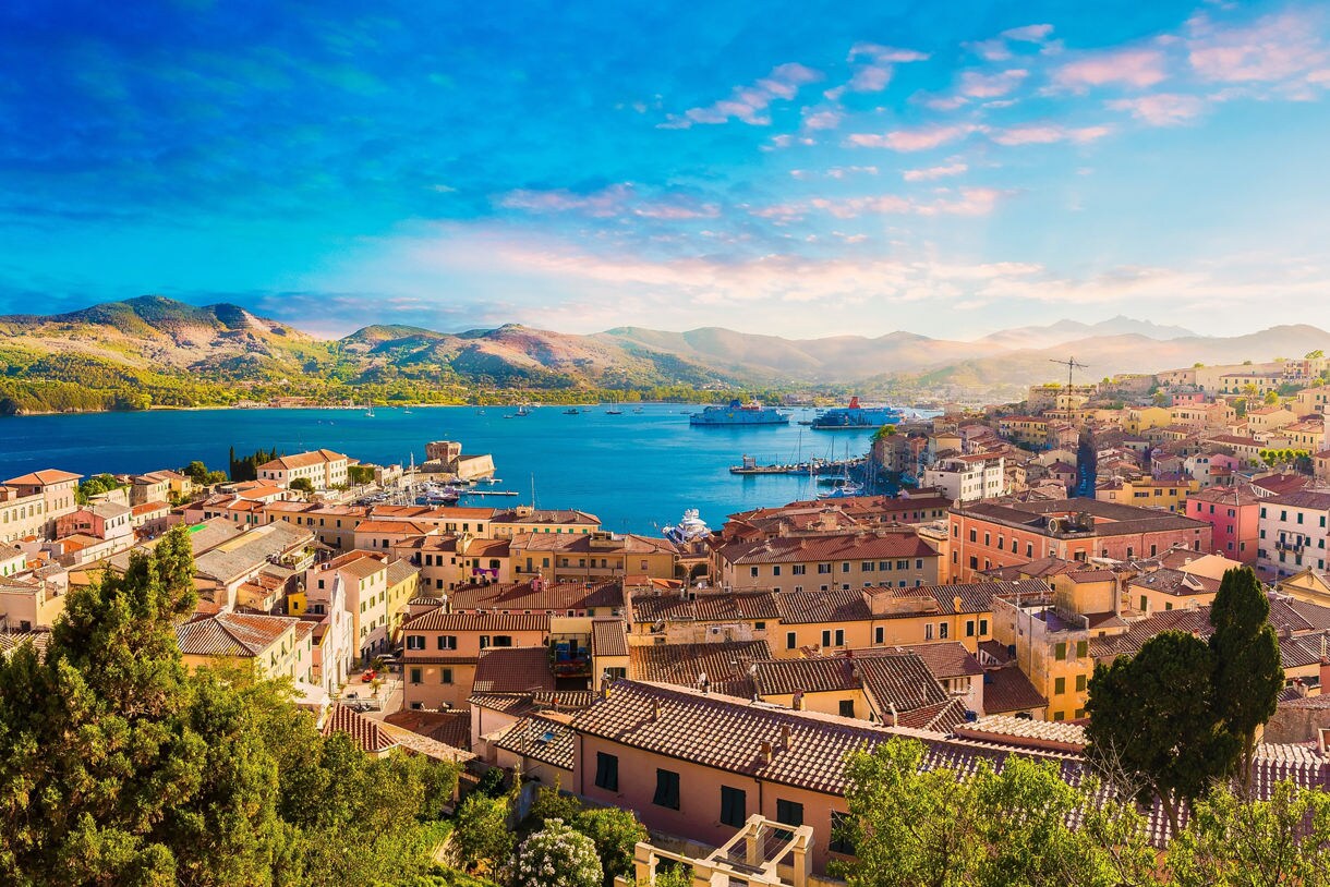 Panoramic view of Portoferraio on Elba Island featuring terracotta rooftops, a bright blue harbor dotted with ferries and sailboats and rolling green mountains under a colorful sky.