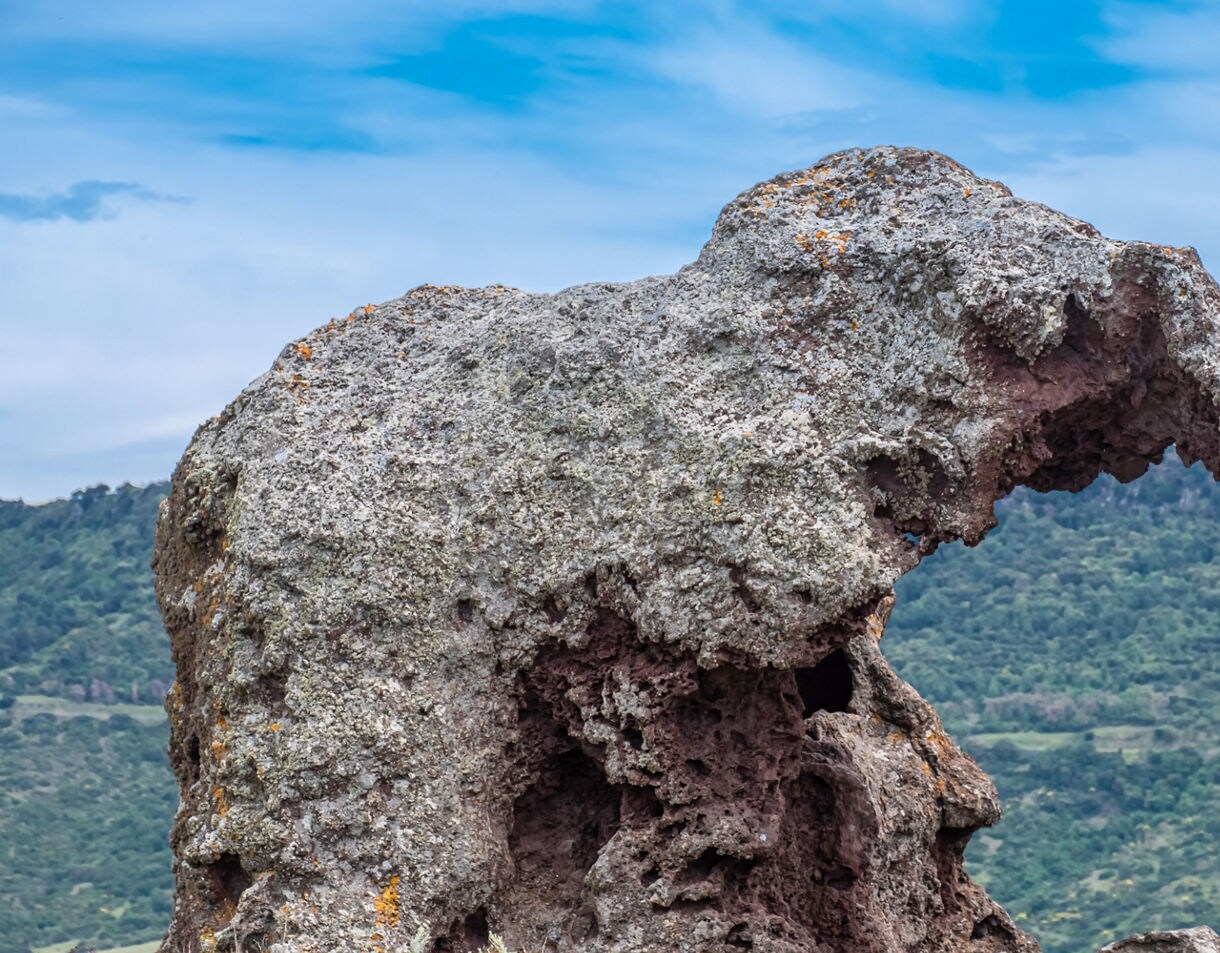 Weathered rock formation resembling an elephant against green hills and a cloudy sky.