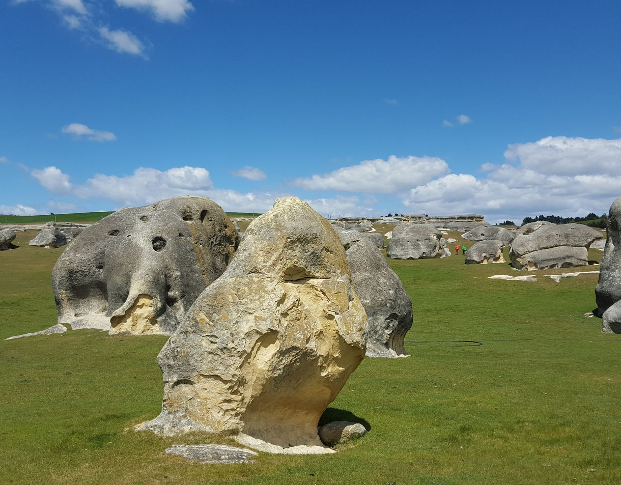 Sunlit limestone boulders scattered across green grasslands, resembling animal shapes beneath a blue sky with scattered clouds.