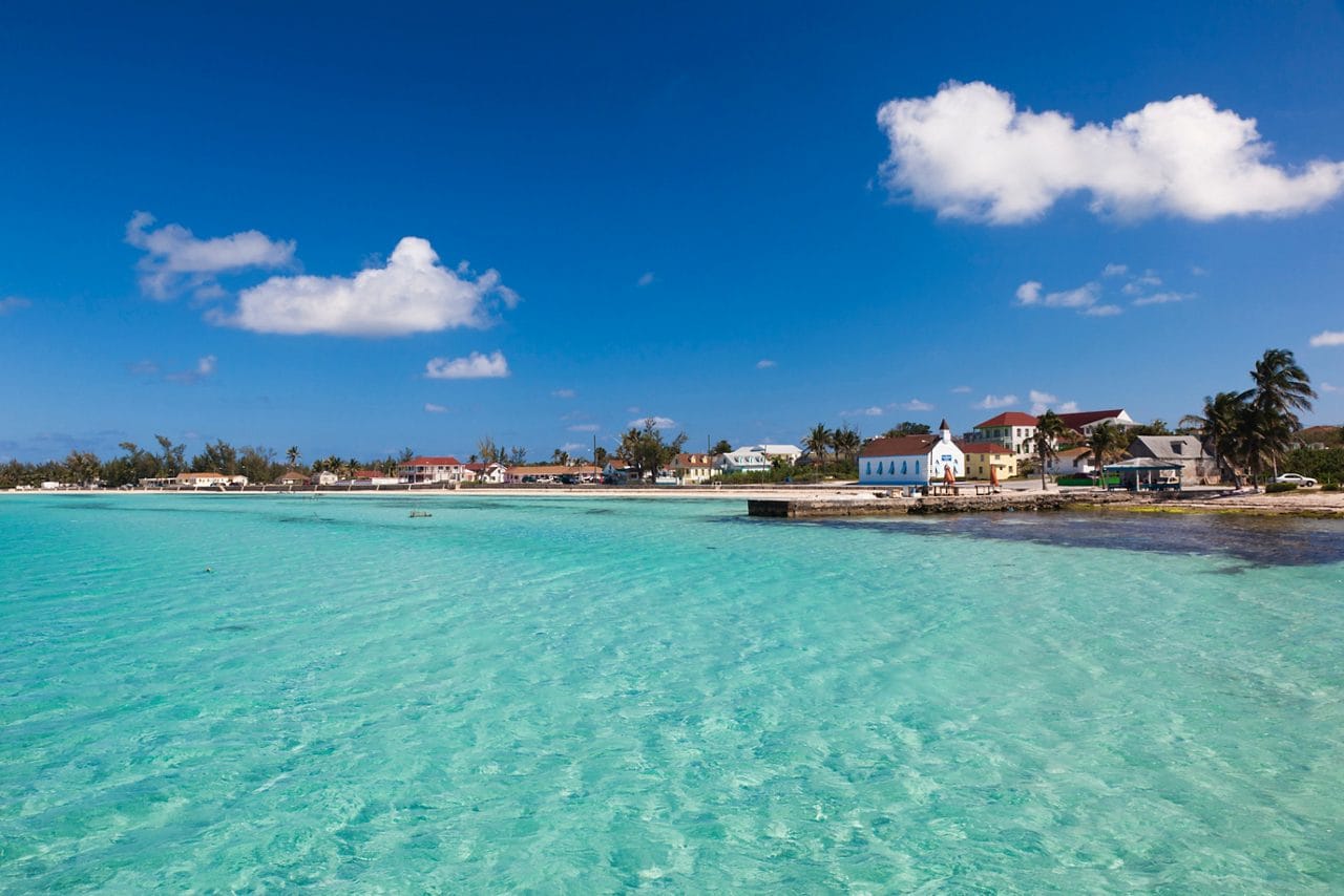 A view of the pristine, shallow waters of Eleuthera Island, Bahamas, with colorful seaside buildings and a clear, sunny sky. 