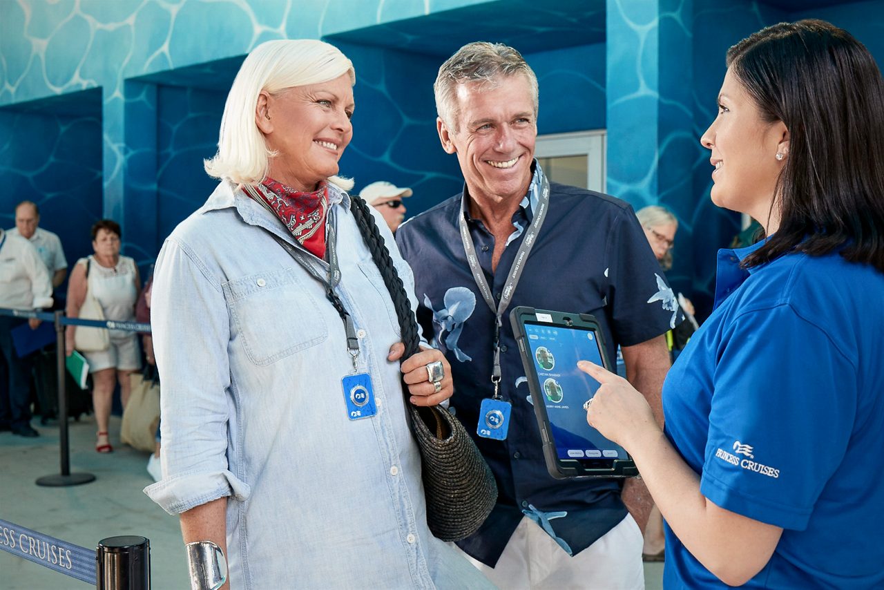 A crew member shows a tablet to two smiling passengers wearing lanyards at an embarkation checkpoint.