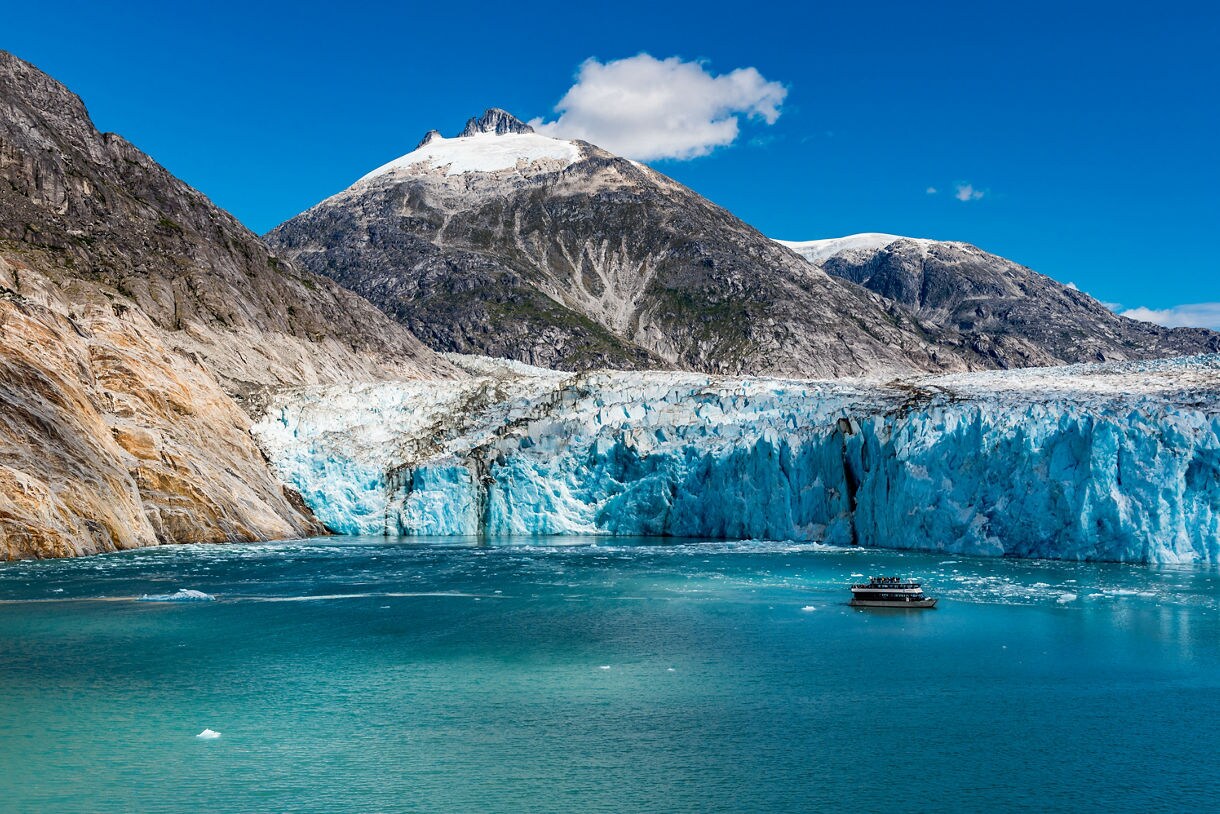A boat floats in front of the massive Dawes Glacier in Alaska, surrounded by towering mountains with snow-covered peaks and bright blue skies.