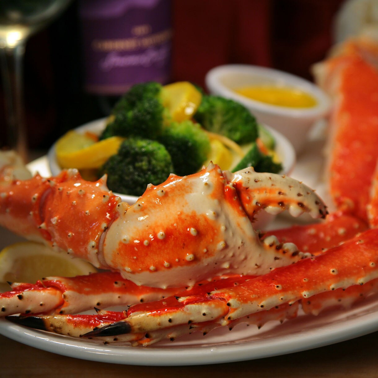 A plate of vibrant king crab legs with a side of broccoli and lemon wedges, accompanied by a glass of white wine in the background.