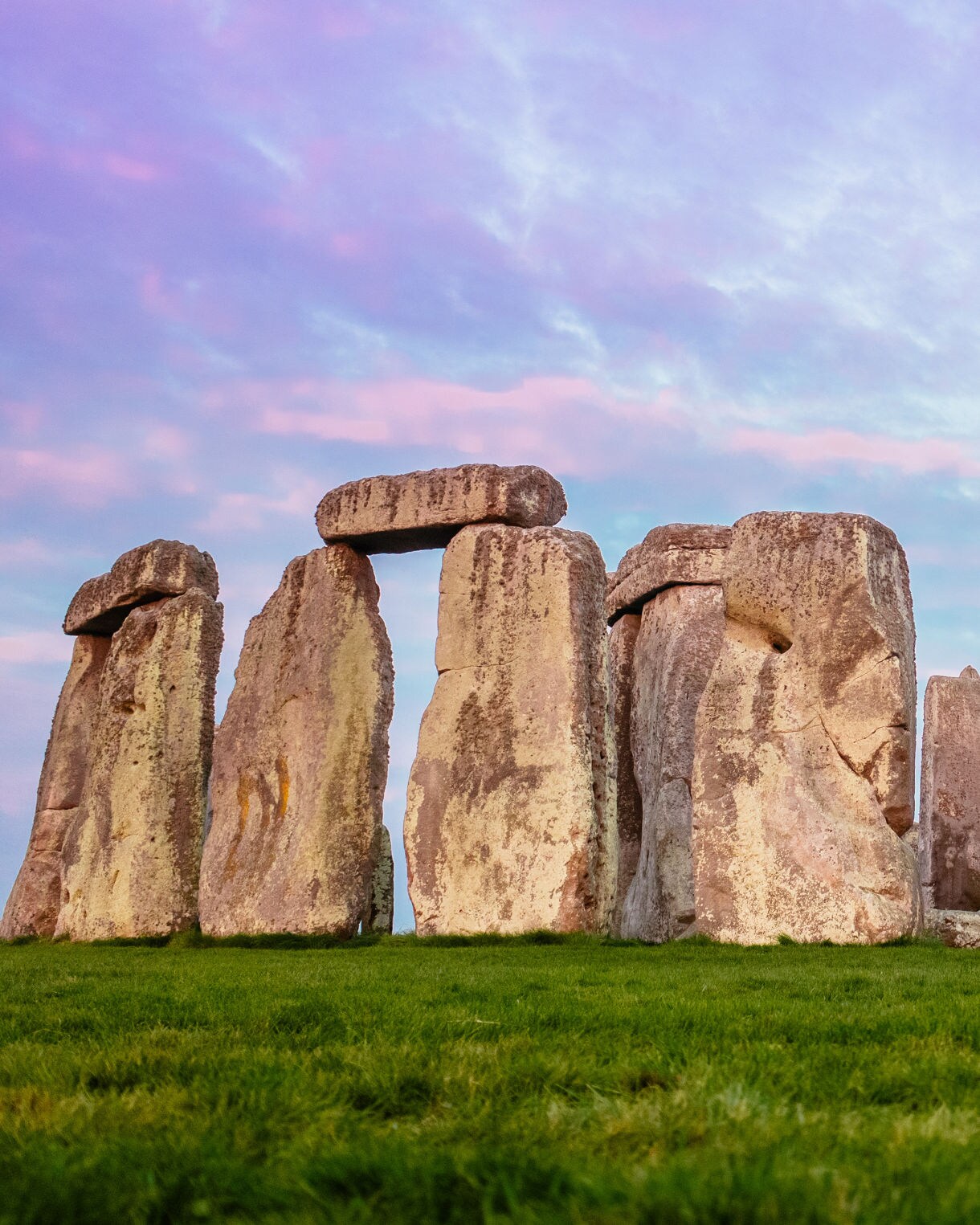 A close-up view of Stonehenge, the iconic prehistoric monument in England, featuring large standing stones arranged in a circular formation. The ancient stones stand tall on a lush green field, with a beautiful sky in the background, colored with soft hues of pink and purple as the sun sets.