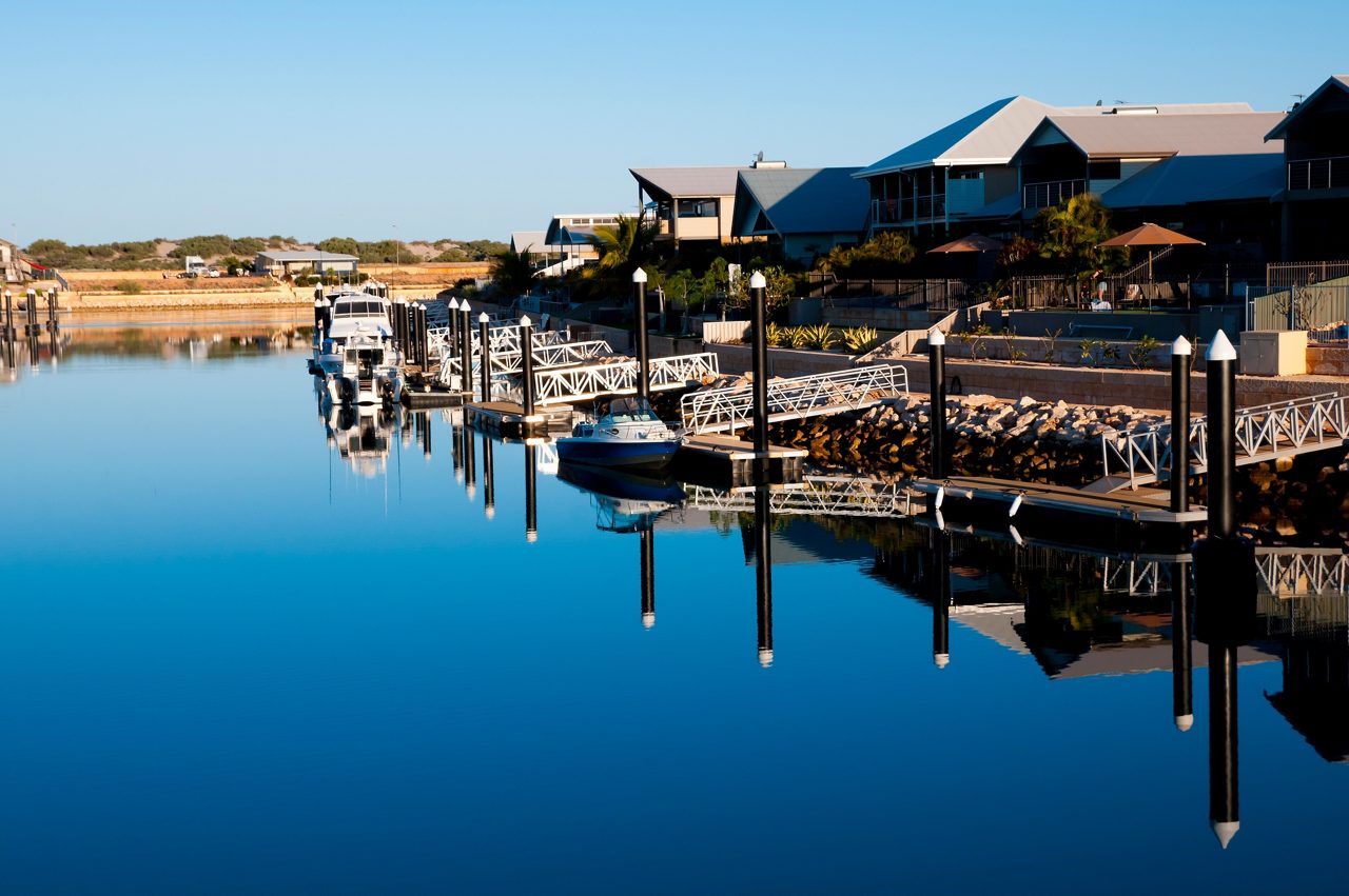 A quiet marina with boats moored at white docks, modern houses lining the waterfront and still blue water reflecting everything clearly.