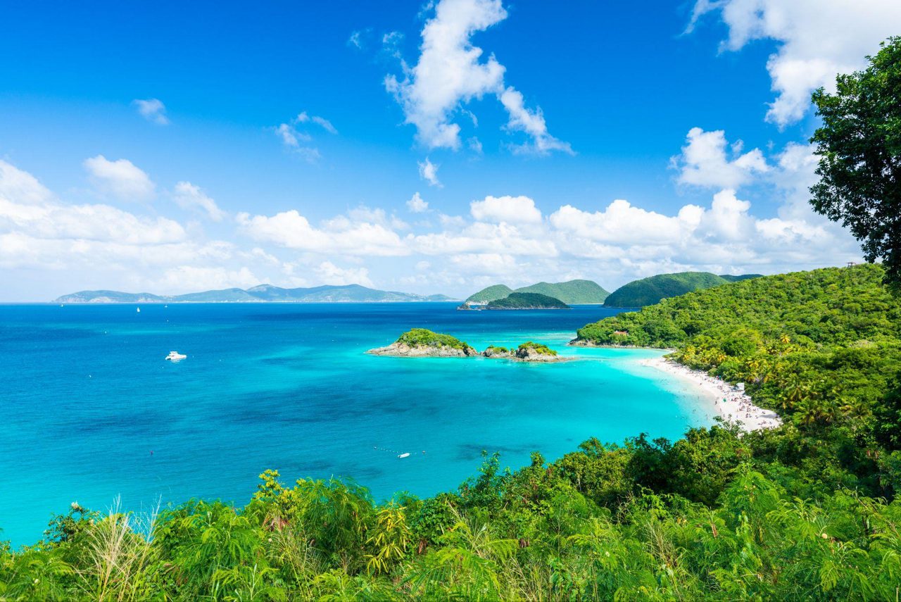 Aerial view of Trunk Bay on St. John, showcasing a white sand beach, turquoise water, offshore islet, dense tropical forest and distant islands under a bright blue sky with scattered clouds.