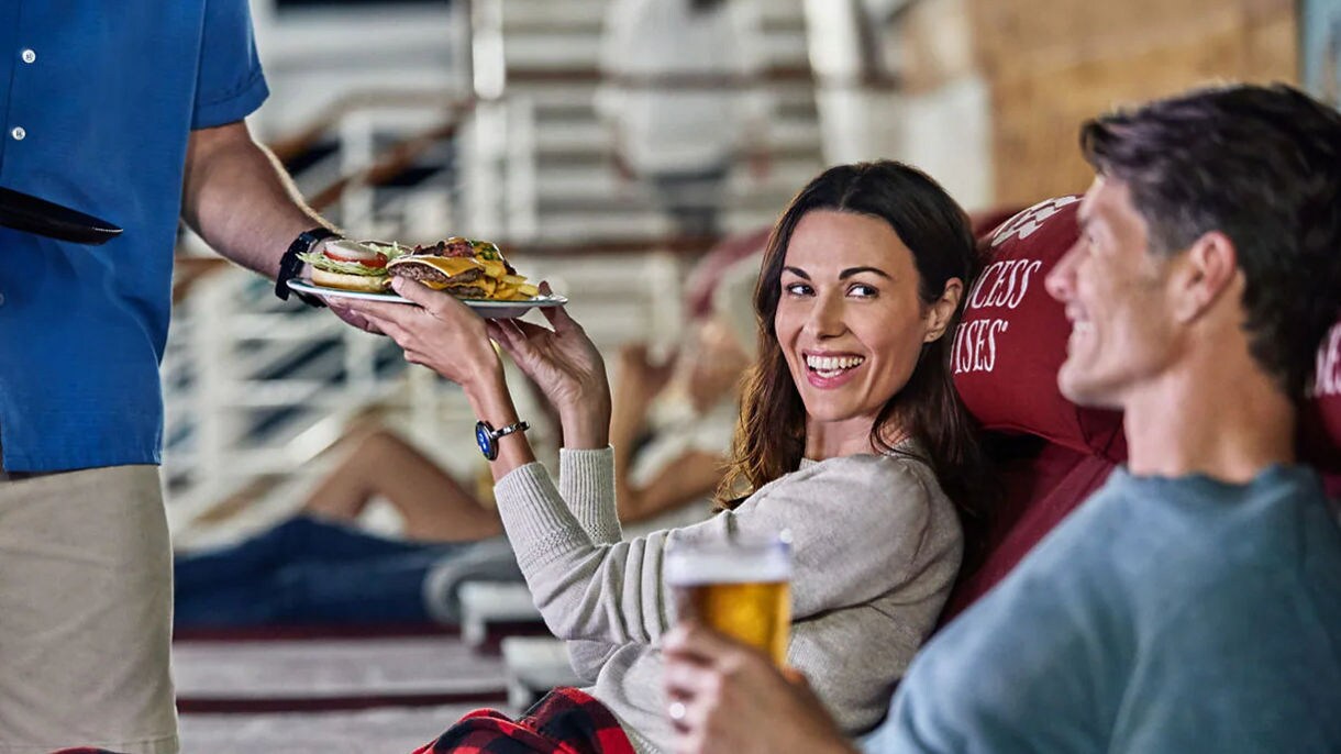 Two smiling adults holding food and beverages on an outdoor cruise ship deck with other passengers visible in the background.