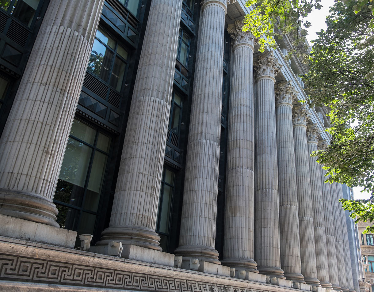 Close-up view of a neoclassical building façade with tall Corinthian columns and decorative stonework partially shaded by leafy trees.