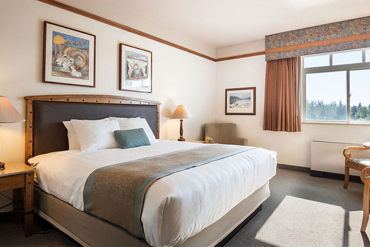 Clean hotel room with white bedding, teal accent pillows, bedside lamp, and framed Alaska artwork on walls.