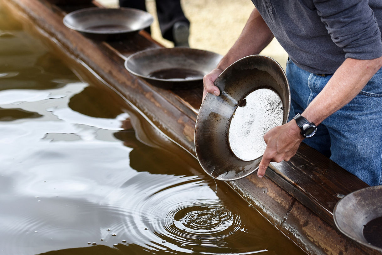 Hands pouring white starter from a pan into a wooden water-filled trough at an outdoor baking demonstration.