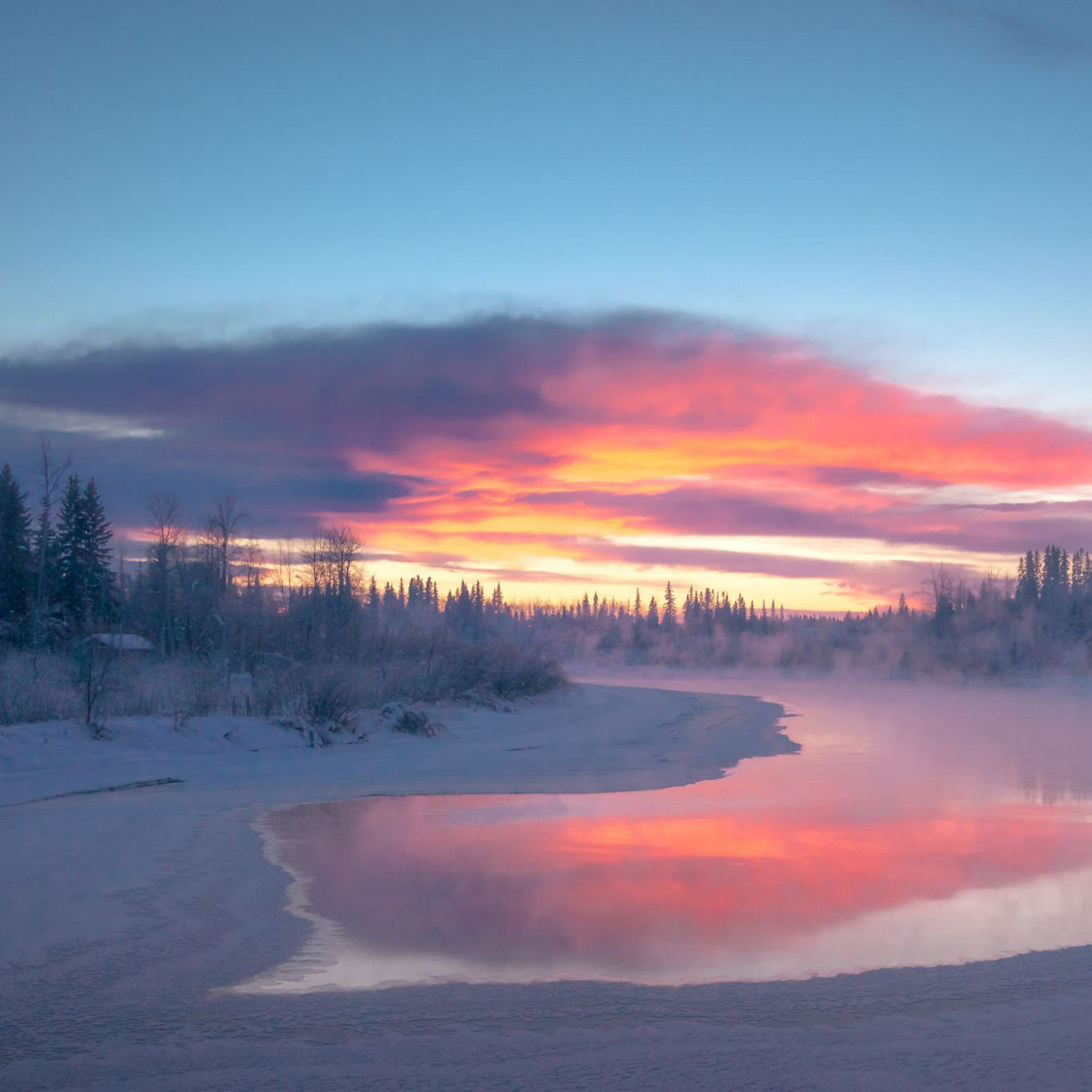 Winter landscape showing a river winding through snow-covered evergreen trees at sunrise, with dramatic pink and orange clouds reflected in the calm water and mist rising from the surface.