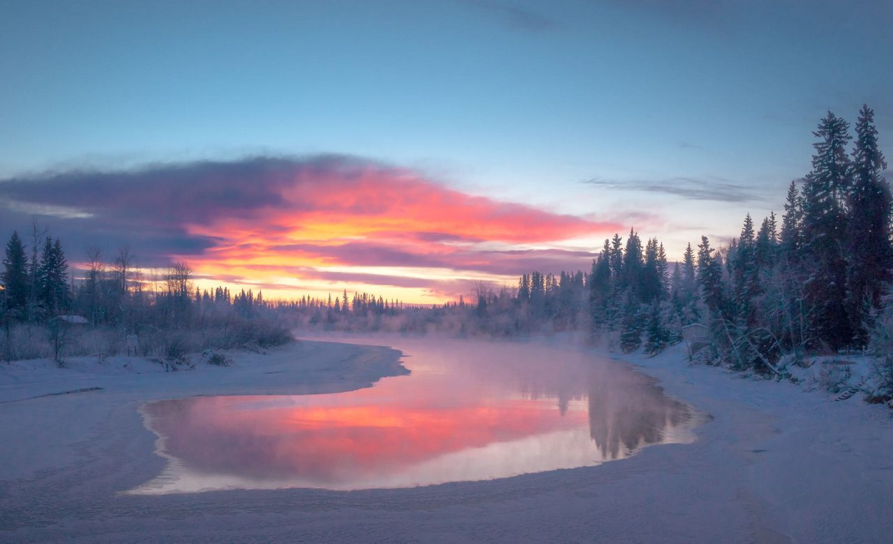 Winter landscape showing a river winding through snow-covered evergreen trees at sunrise, with dramatic pink and orange clouds reflected in the calm water and mist rising from the surface.