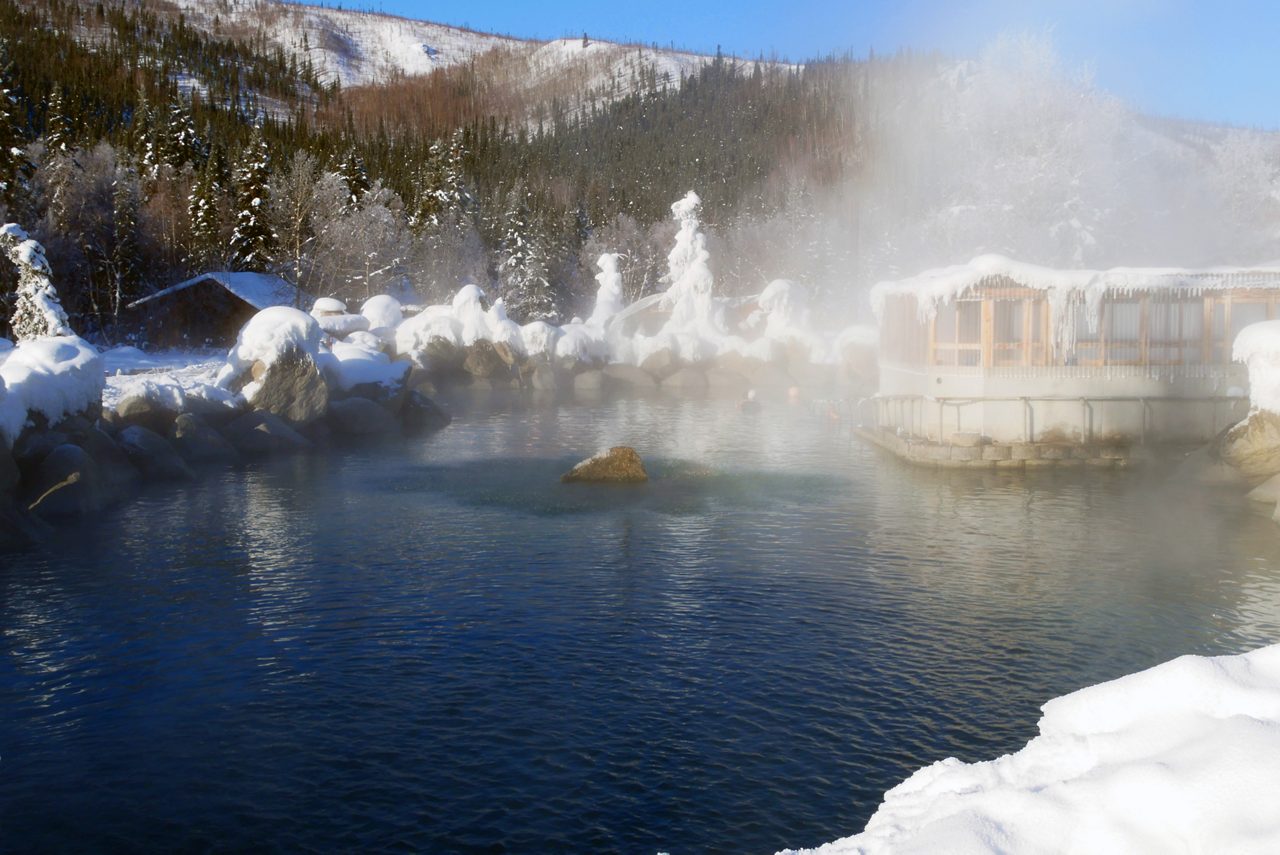 Steamy outdoor hot springs surrounded by snow-covered boulders and icicle-lined buildings, nestled in a forested mountain area near Fairbanks, Alaska.