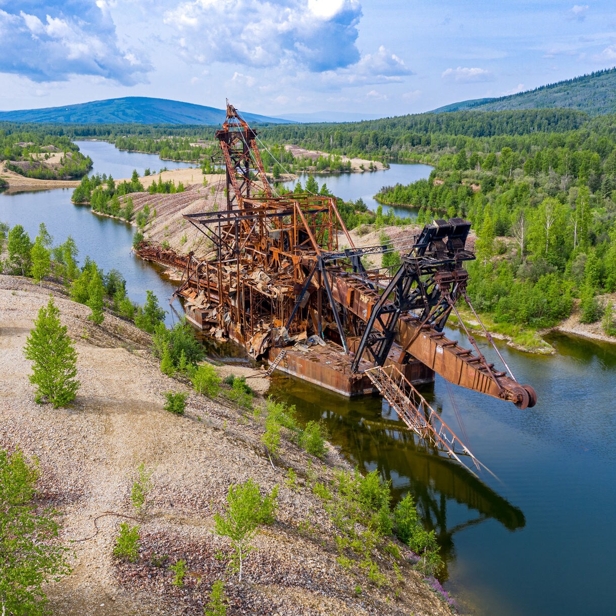 Aerial view of an abandoned, rusted gold dredge surrounded by rocky terrain, green trees, and winding water channels in the hills near Fairbanks, Alaska.
