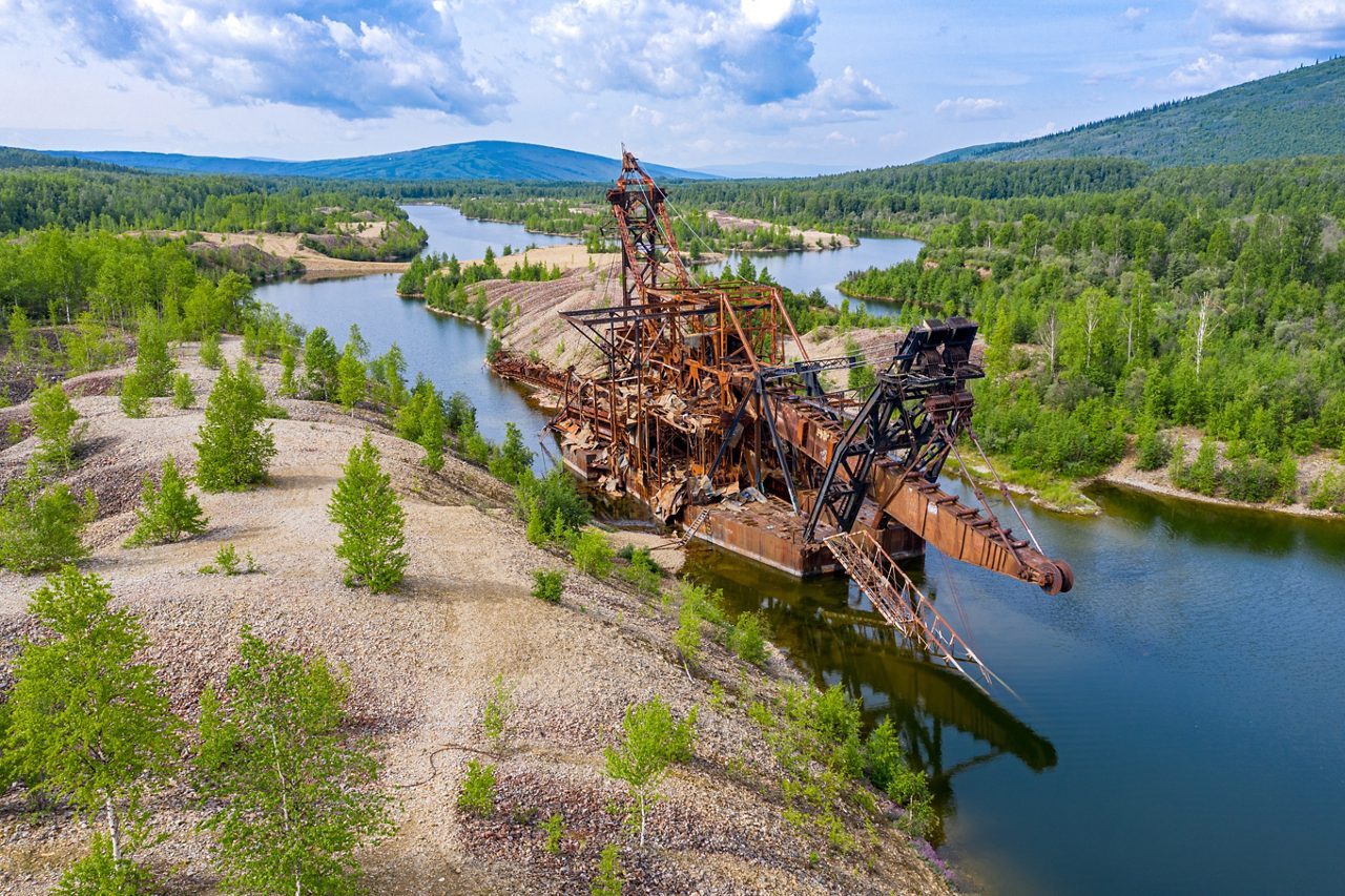 Aerial view of an abandoned, rusted gold dredge surrounded by rocky terrain, green trees, and winding water channels in the hills near Fairbanks, Alaska.