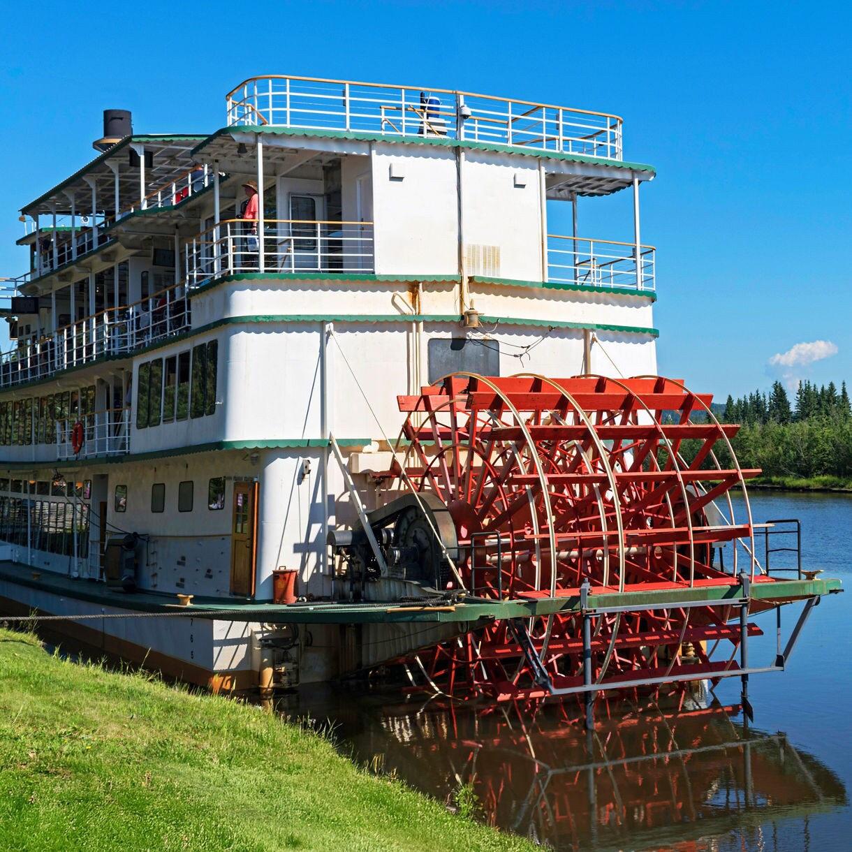 Restored Canadian Pacific sternwheeler docked beside a lake, with a red paddlewheel, wooden walkway, picnic tables, and forested mountains in the background.