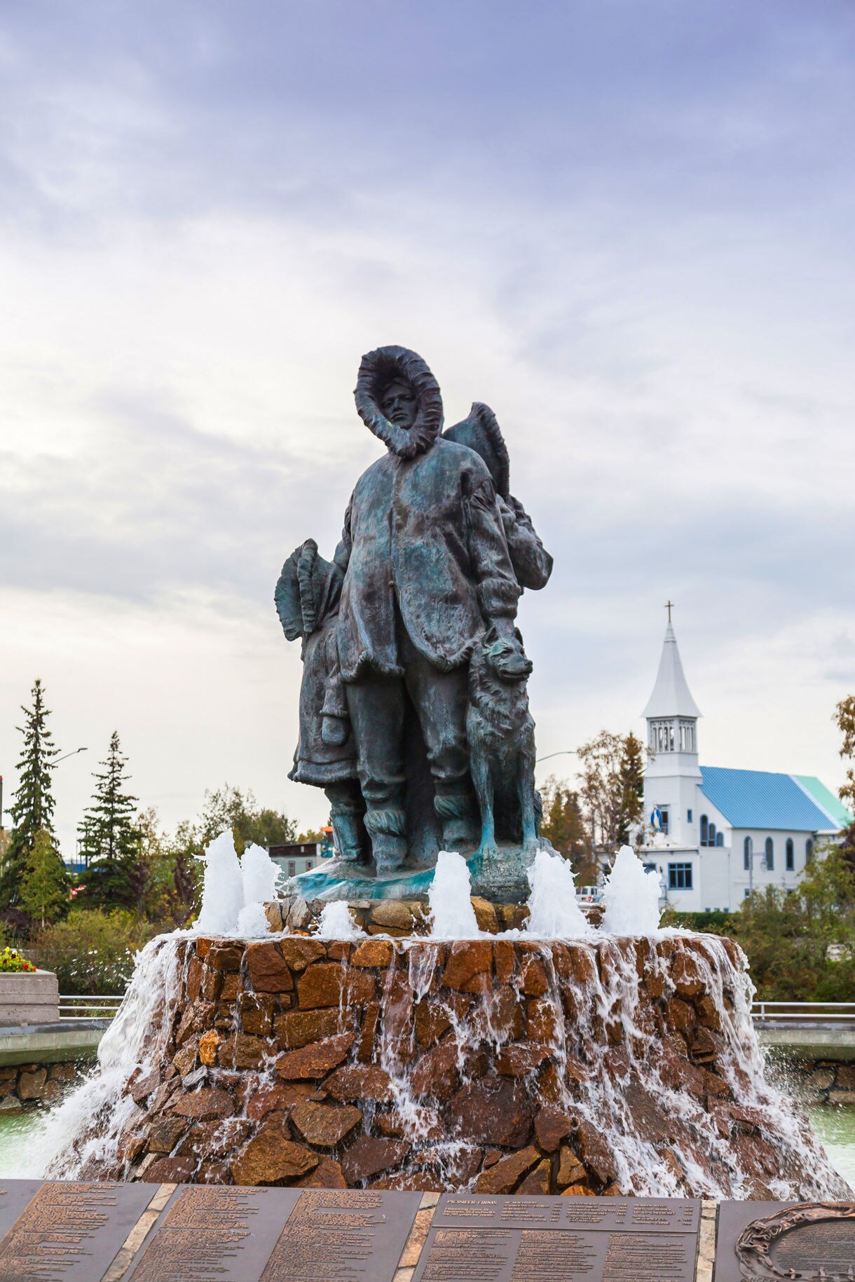 Bronze statue of an Alaska Native family and dog wearing traditional parkas, standing atop a rock fountain with cascading water, with a white church and pine trees in the background.