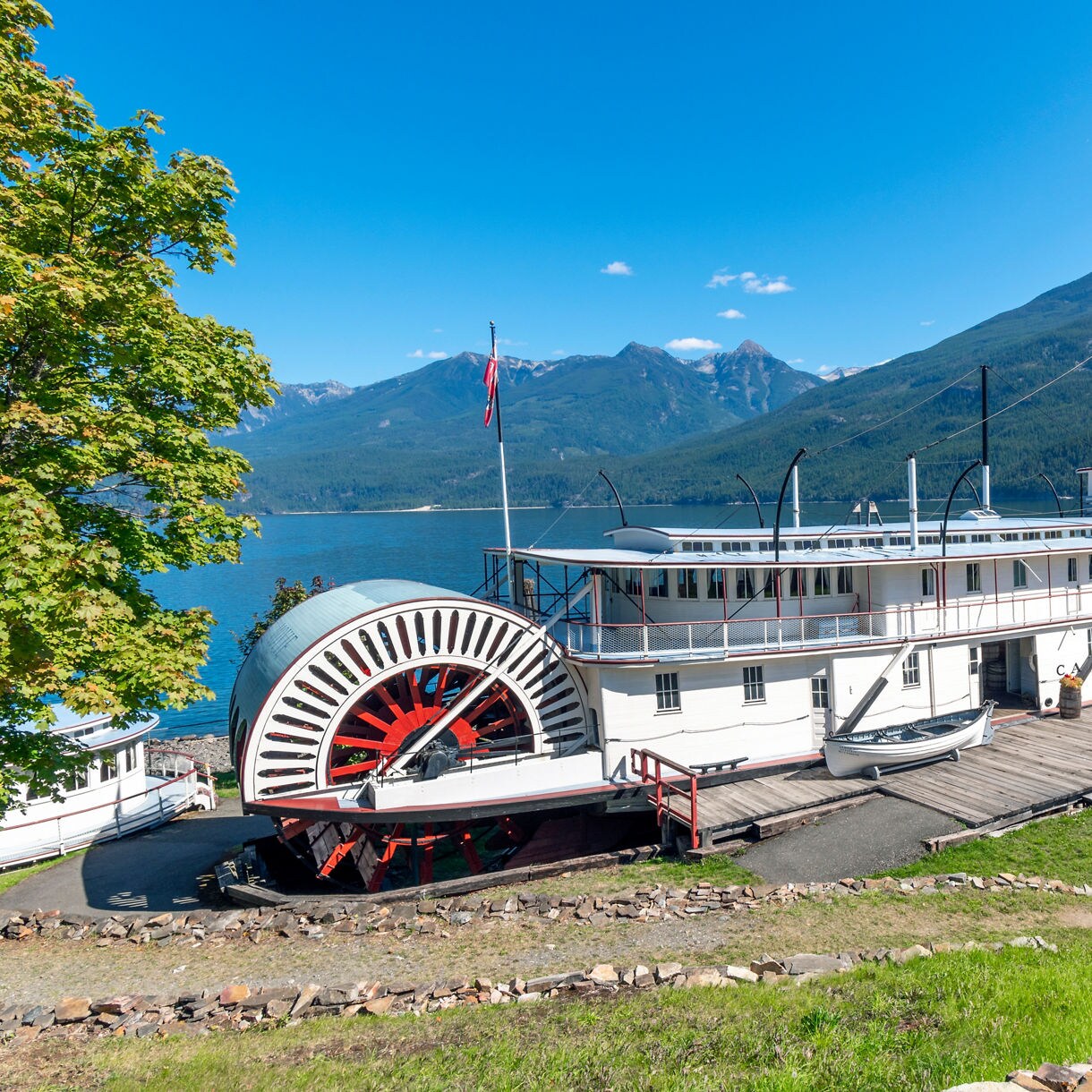 Restored Canadian Pacific sternwheeler docked beside a lake, with a red paddlewheel, wooden walkway, picnic tables, and forested mountains in the background