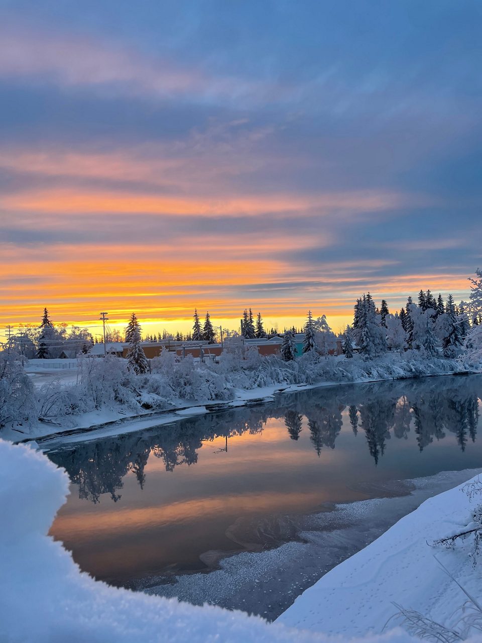 Vibrant orange and purple sunset over a snowy Fairbanks landscape, with snow-laden trees and buildings reflected in the calm, partially frozen river below.