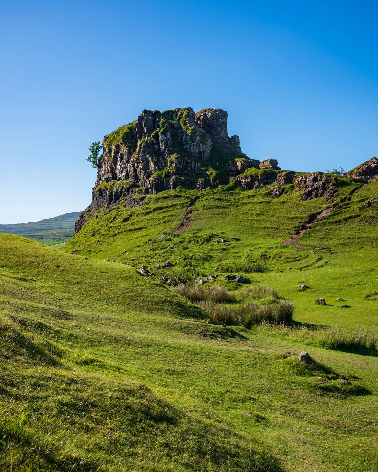 Sunlit grassy hills surrounding a rocky, flat-topped outcrop at the Fairy Glen on the Isle of Skye, with bright green slopes, scattered stones and a clear blue sky overhead.