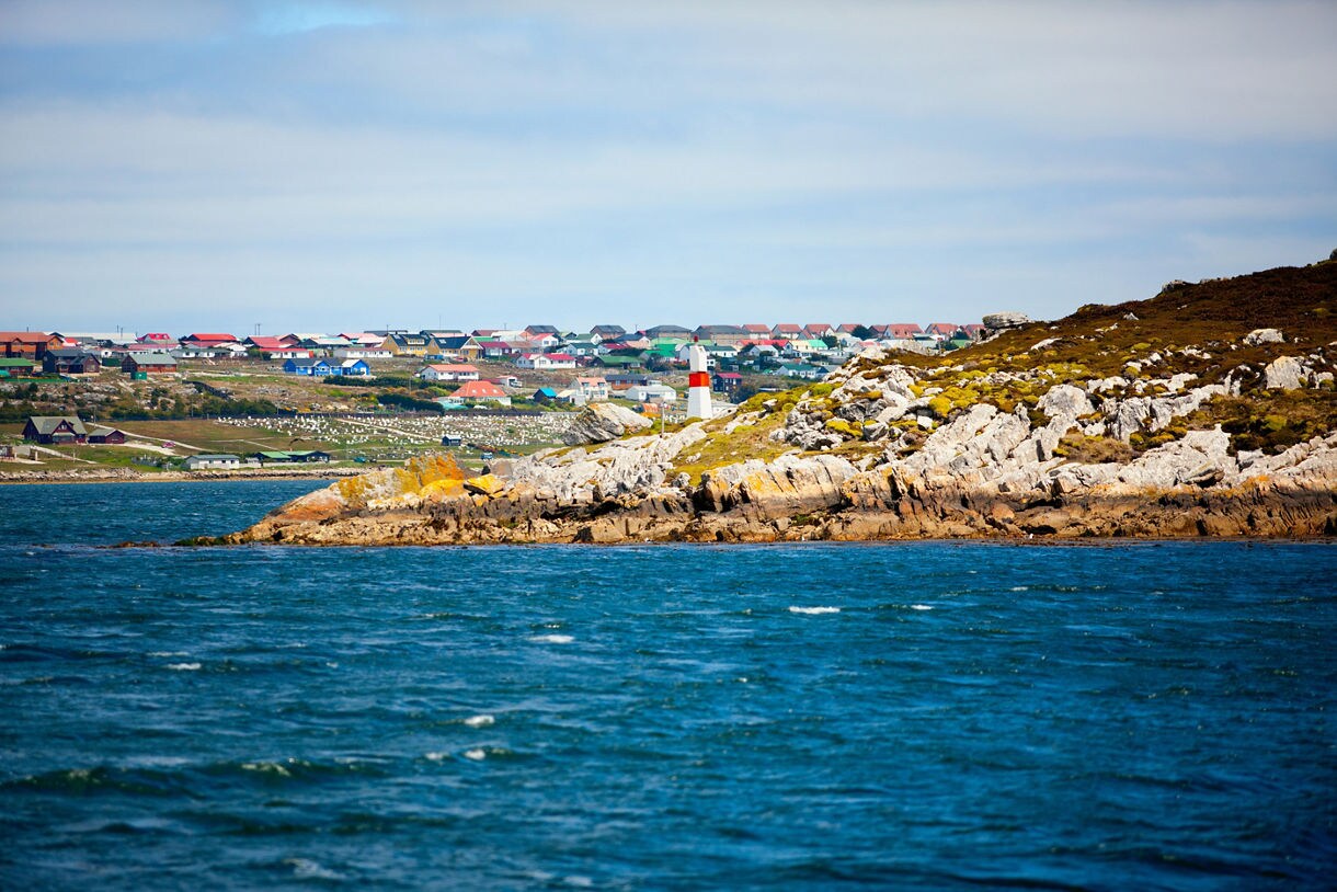 Rocky shoreline with a small red-and-white lighthouse in front of a distant town of colorful houses beside blue choppy water.