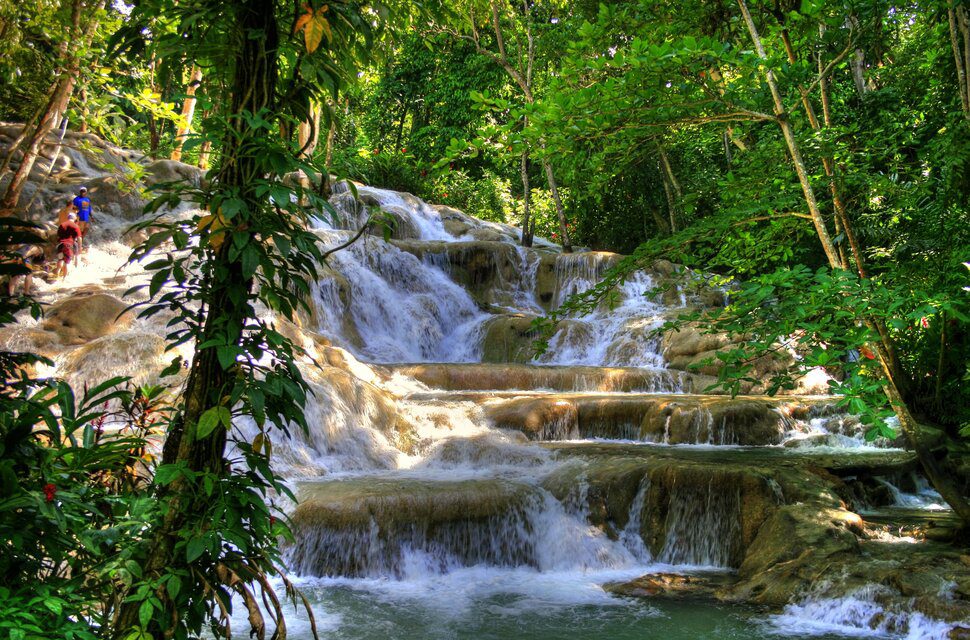 Dunn’s River Falls in Jamaica featuring terraced limestone rock formations, clear rushing water and dense green foliage with visitors climbing the waterfall.