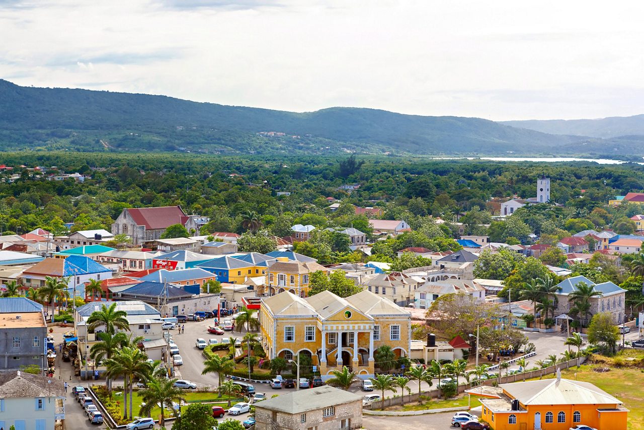 Aerial view of Falmouth, Jamaica featuring colonial-era Georgian buildings, colorful homes, a yellow courthouse, a white church tower and green hills stretching toward the horizon.