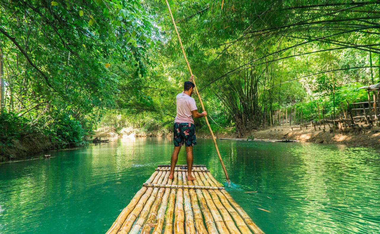 A man steering a bamboo raft along the calm Martha Brae River, surrounded by dense tropical greenery and arched bamboo trees forming a natural canopy.