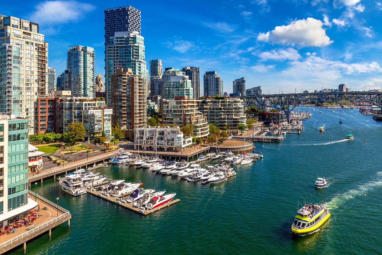 False Creek in Vancouver with modern high-rise buildings, a marina filled with docked boats, a yellow water taxi cruising through the inlet and a bridge spanning the water under a bright blue sky.