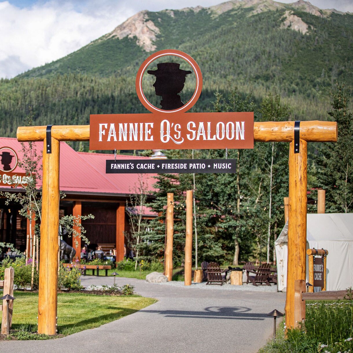 The image shows the entrance to Fannie Q's Saloon, marked by a prominent wooden sign and archway. The saloon building features a rustic design with a red roof, surrounded by lush greenery and colorful flowers. In the background, a scenic mountain rises under a partly cloudy sky, creating a welcoming and picturesque outdoor setting. Visible text includes 'Fannie Q's Saloon' and 'Fannie's Cache • Fireside Patio • Music.'