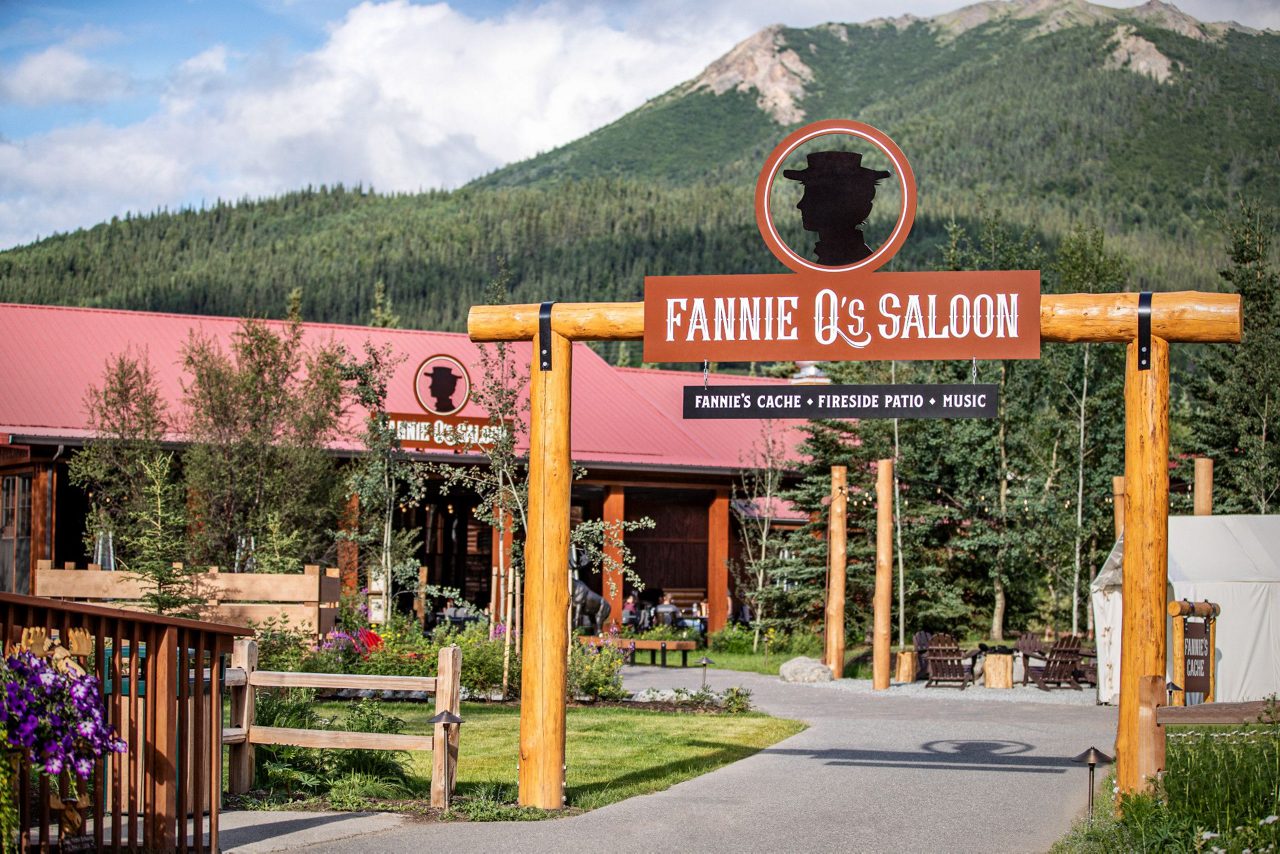 The image shows the entrance to Fannie Q's Saloon, marked by a prominent wooden sign and archway. The saloon building features a rustic design with a red roof, surrounded by lush greenery and colorful flowers. In the background, a scenic mountain rises under a partly cloudy sky, creating a welcoming and picturesque outdoor setting. Visible text includes 'Fannie Q's Saloon' and 'Fannie's Cache • Fireside Patio • Music.'
