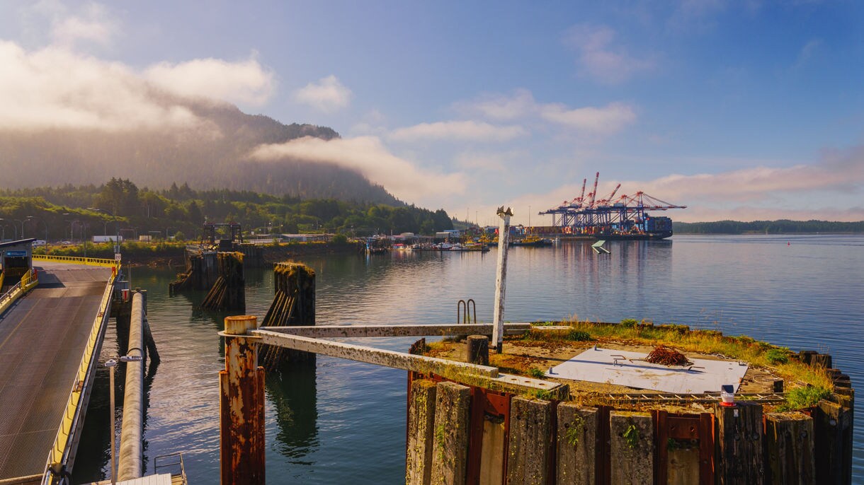 A ferry docked at a pier in Prince Rupert with mountains and clouds in the background.