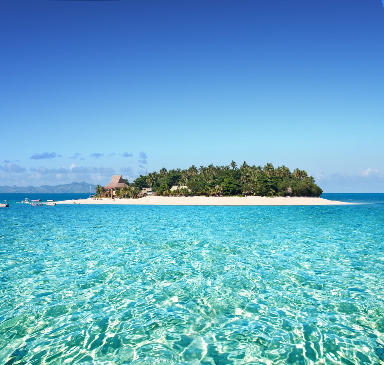 A small tropical island in Fiji, South Pacific, surrounded by vibrant turquoise waters and fringed with lush greenery and white sand beaches, under a bright blue sky. 