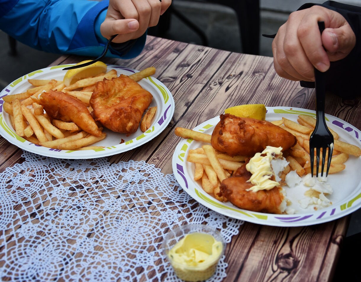 Two plates of golden fried fish with French fries, lemon wedges and mayonnaise on a wooden table.