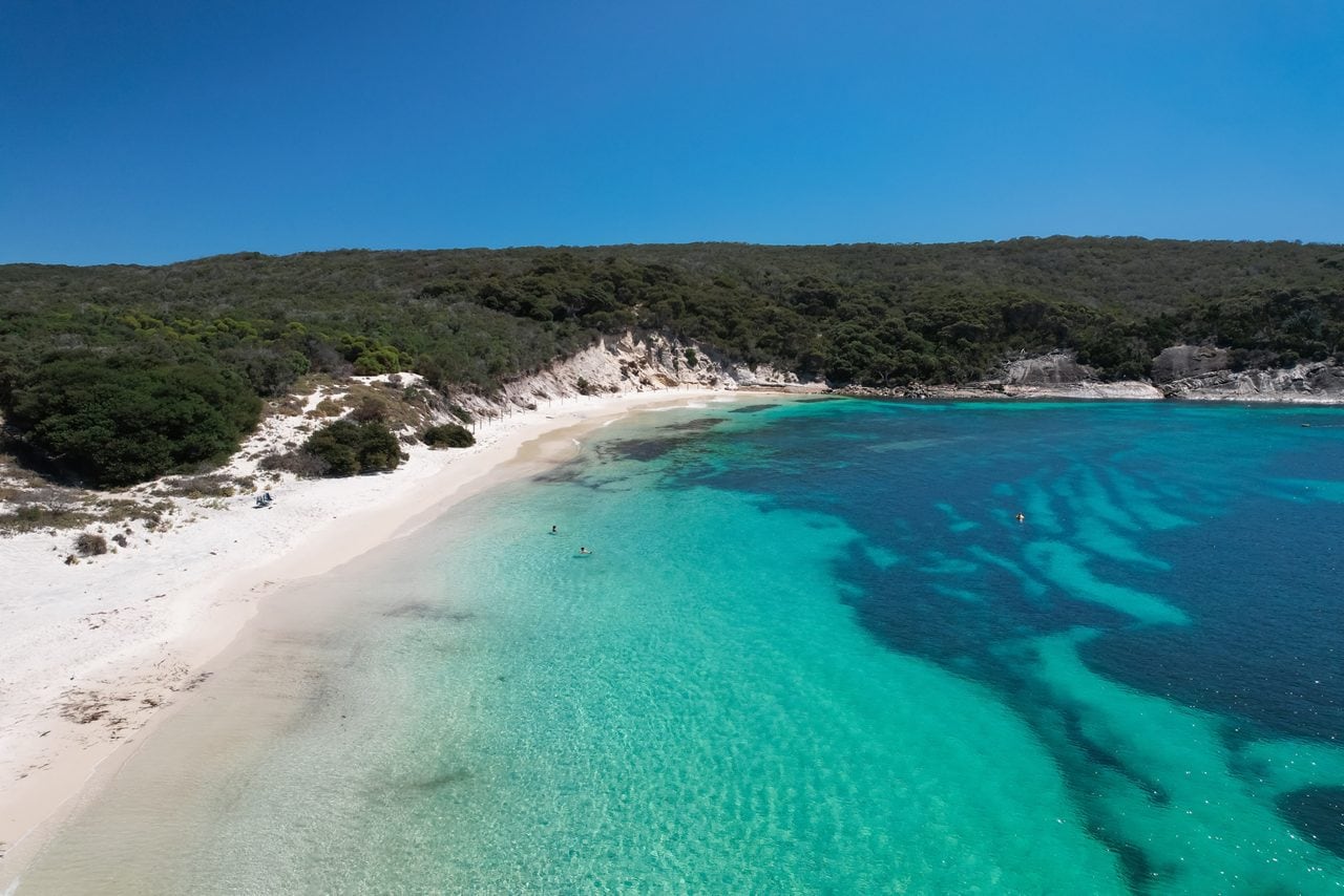 A secluded white-sand beach with turquoise shallows and deeper blue water, bordered by dense green coastal vegetation under a bright blue sky.
