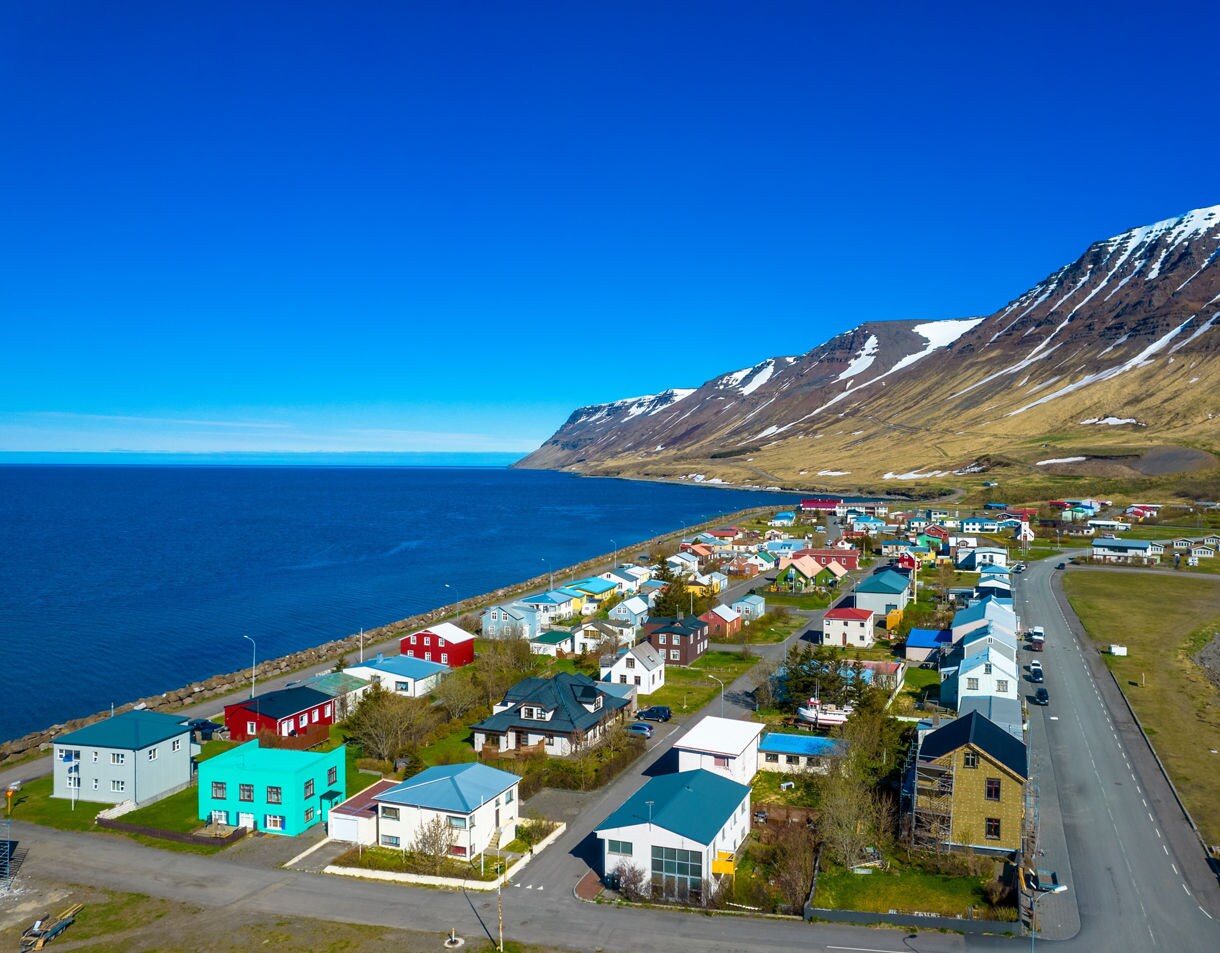 Aerial view of Flateyri village in Iceland with colorful houses along the fjord, mountains with patches of snow in the background and clear blue skies overhead.