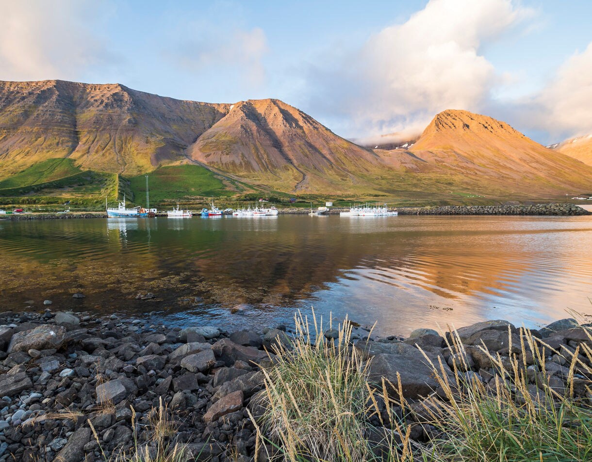Fishing boats moored in Flateyri fjord with rugged mountains glowing in evening sunlight.
