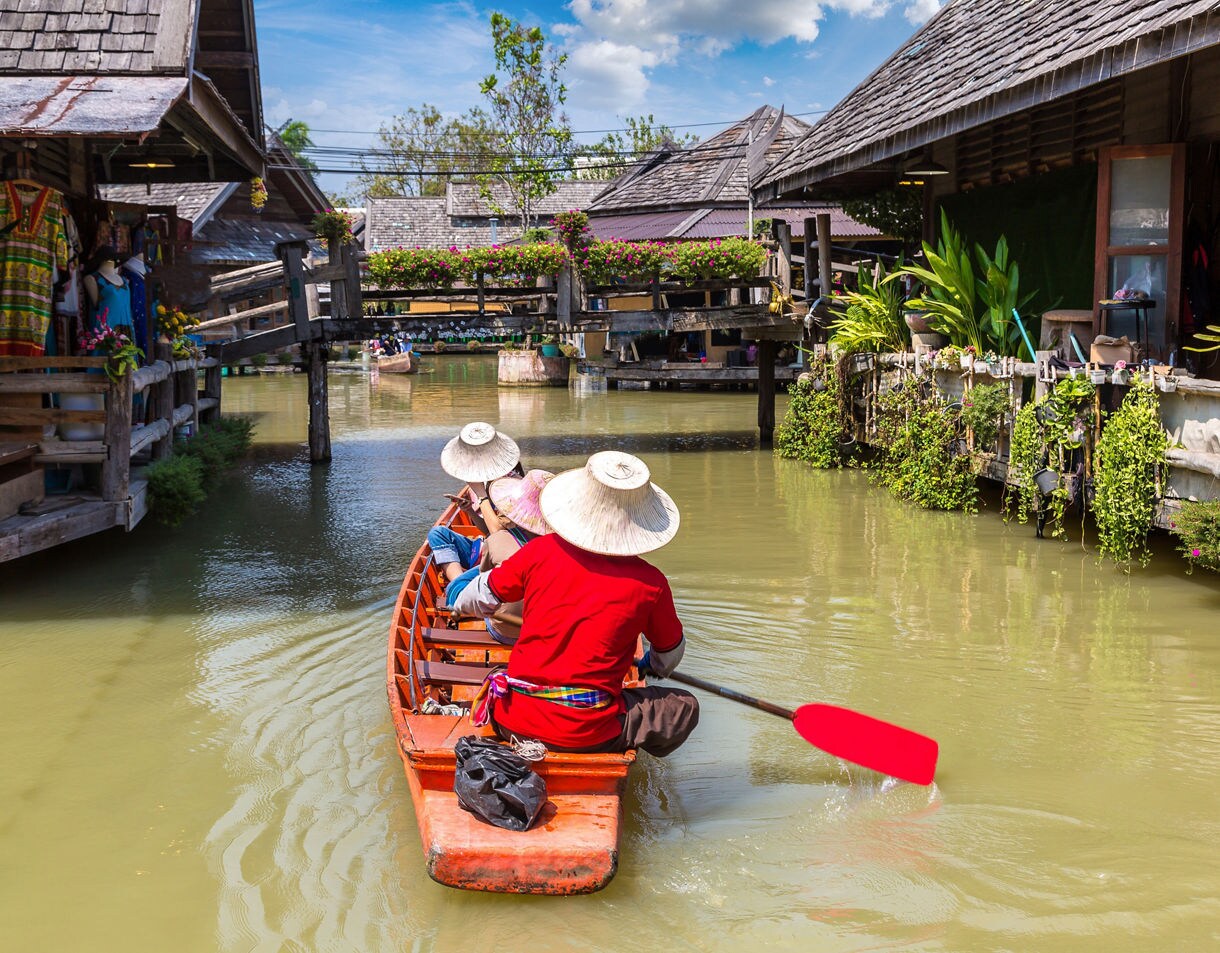 People paddling a wooden boat along a canal lined with traditional stilt houses, market stalls and plants at a floating market.