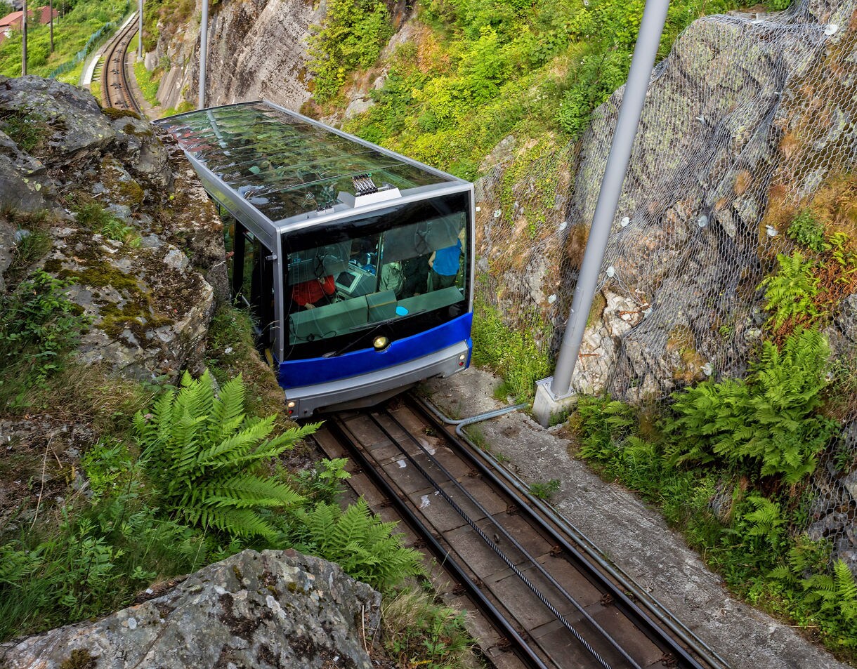 A blue and silver Fløibanen funicular car travels between rocky cliffs covered in greenery, heading up to Mount Fløyen in Bergen, Norway.