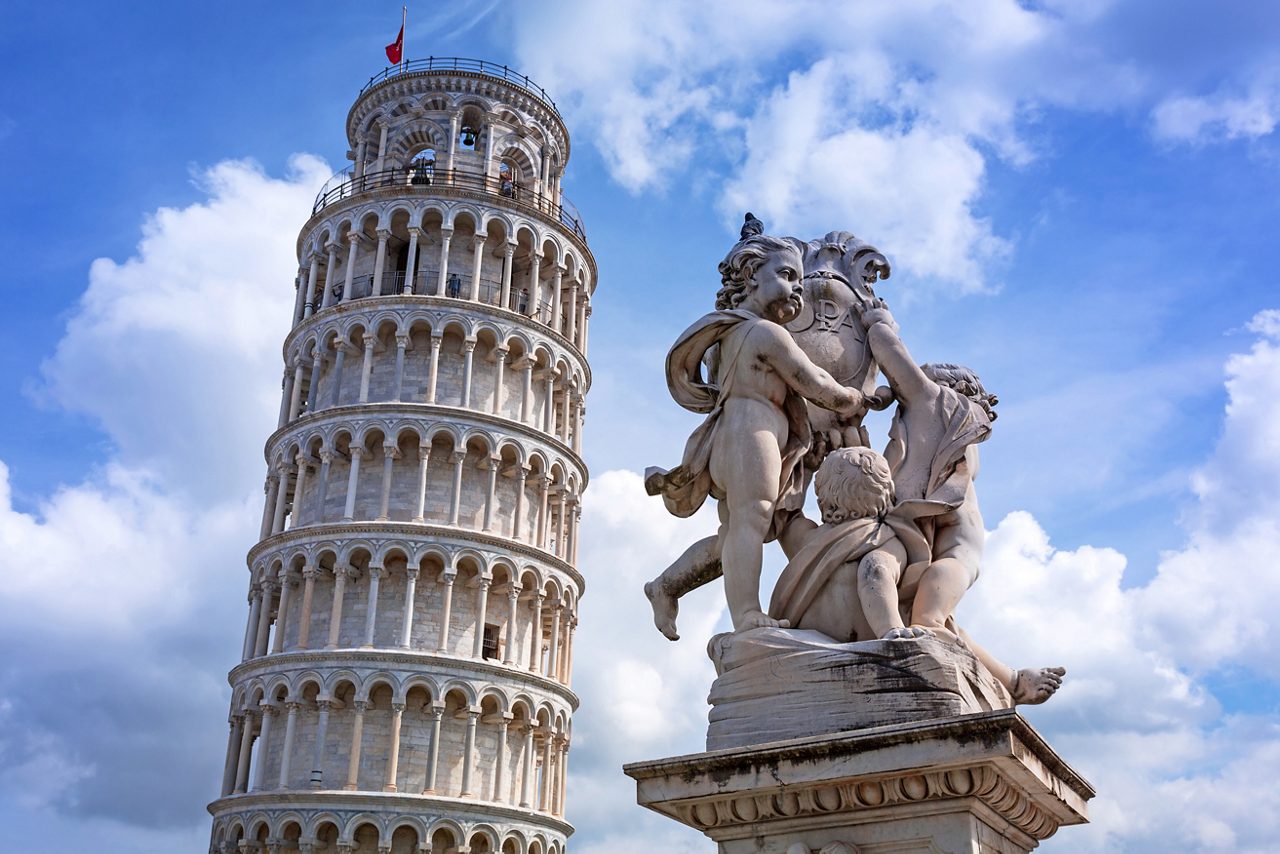 The Leaning Tower of Pisa in Italy, standing at an angle against a bright blue sky, with a detailed marble statue of cherubs in the foreground.