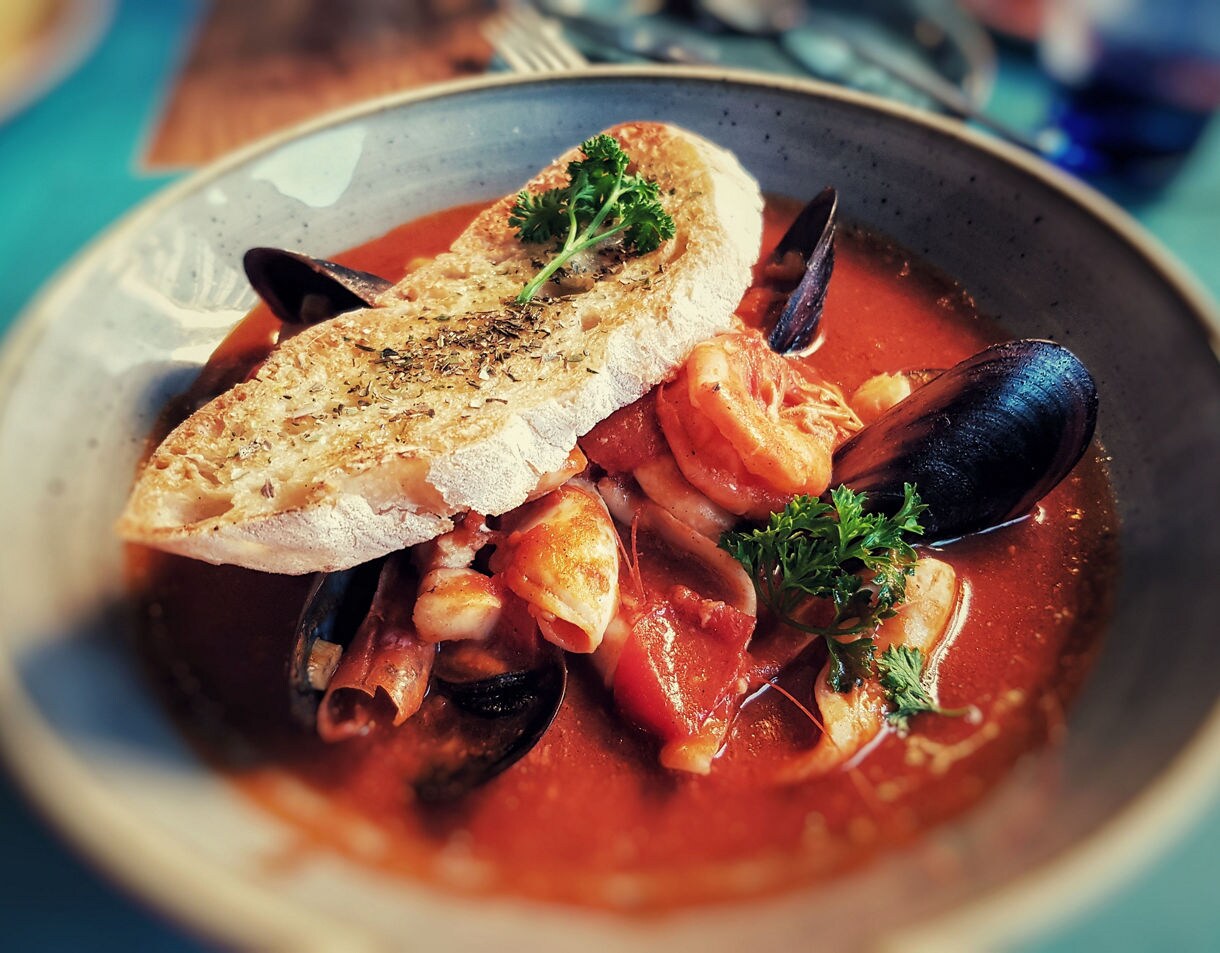 Close-up of a bowl of tomato-based seafood stew with shrimp, mussels and fish, garnished with parsley and served with a slice of toasted bread on top.